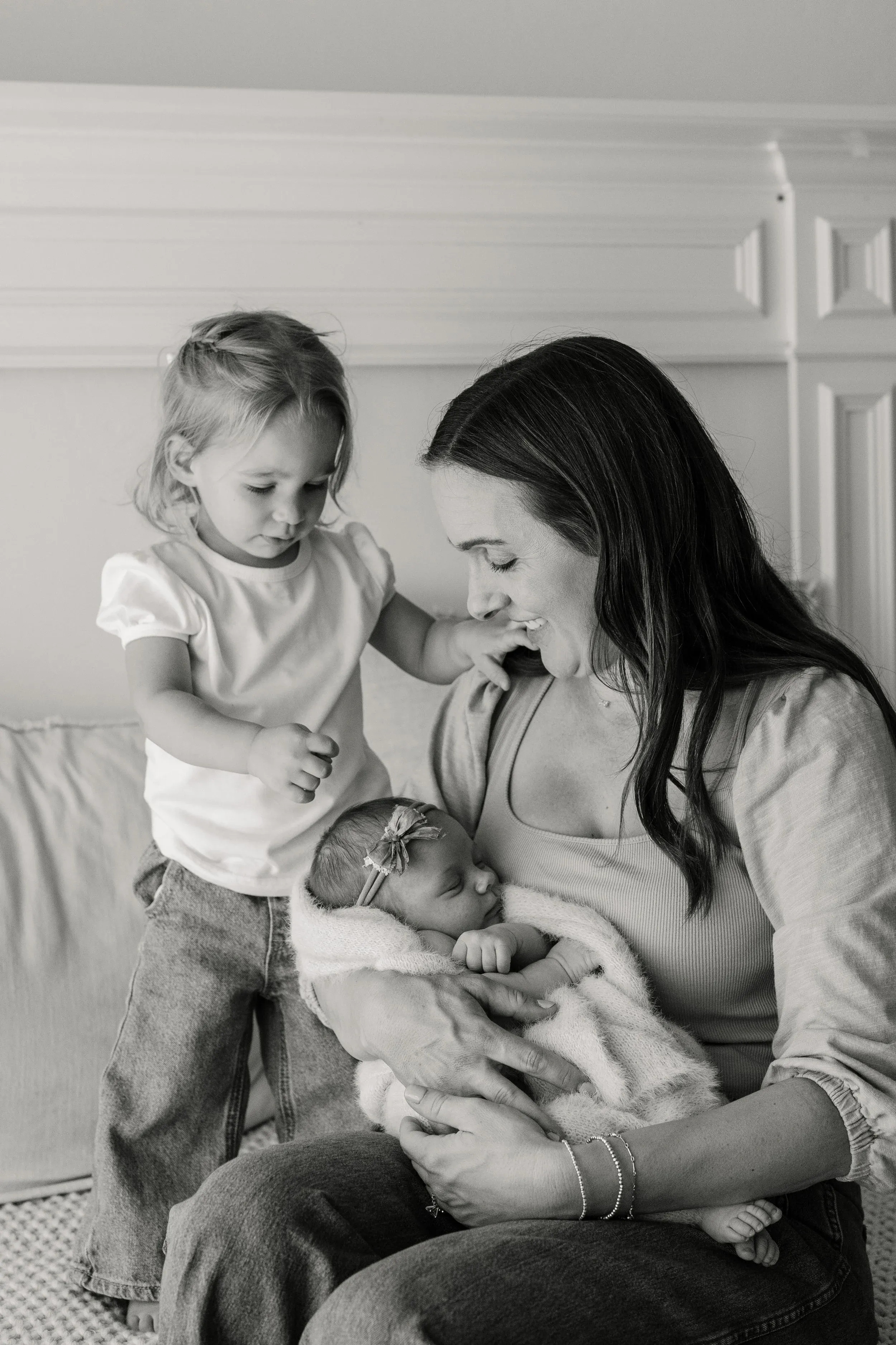Black and white photo of Caitlin Richardson smiling down at a newborn in her arms while a toddler leans on her shoulder