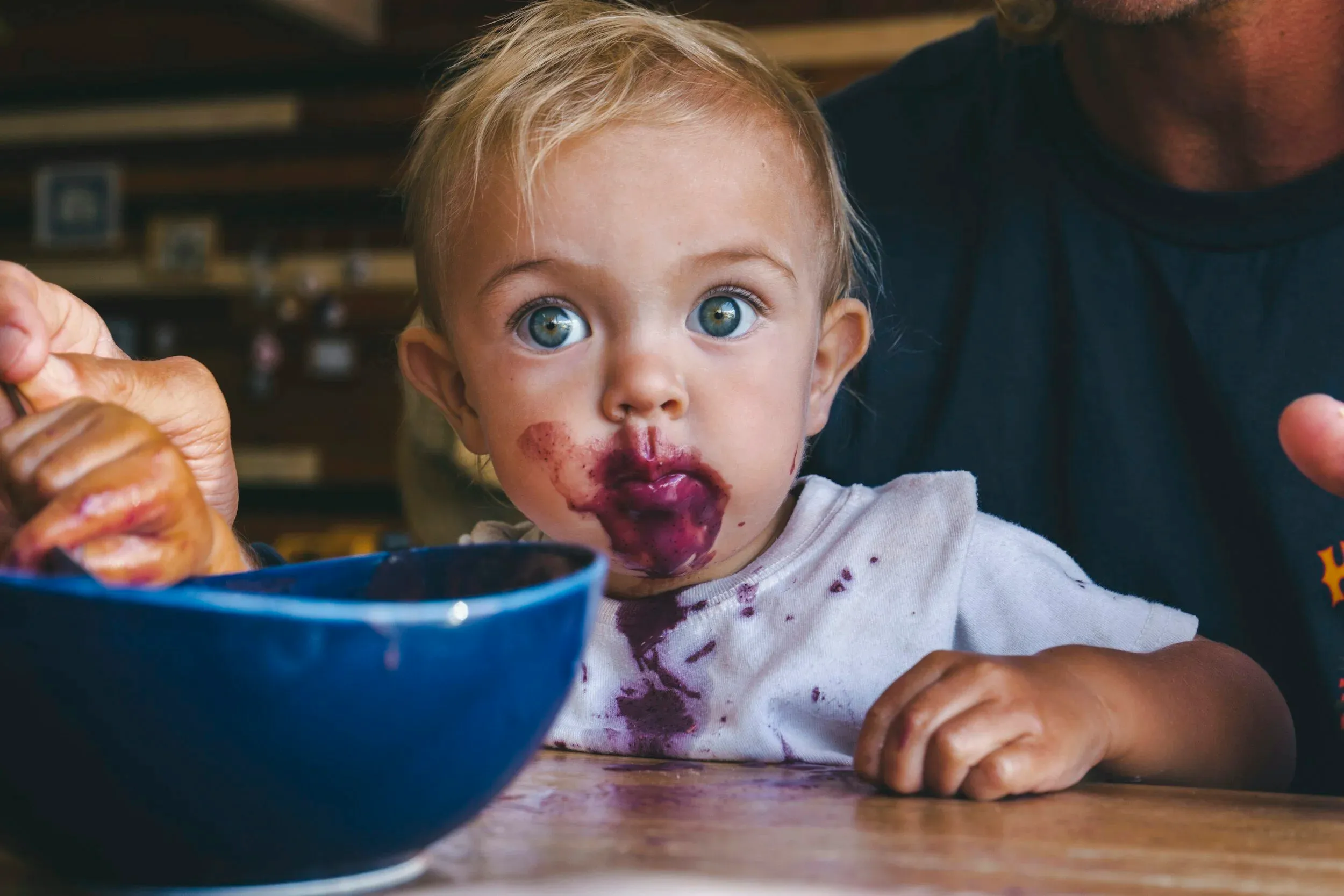 A toddler with a berry-stained face eating from a bowl while a parent watches from behind