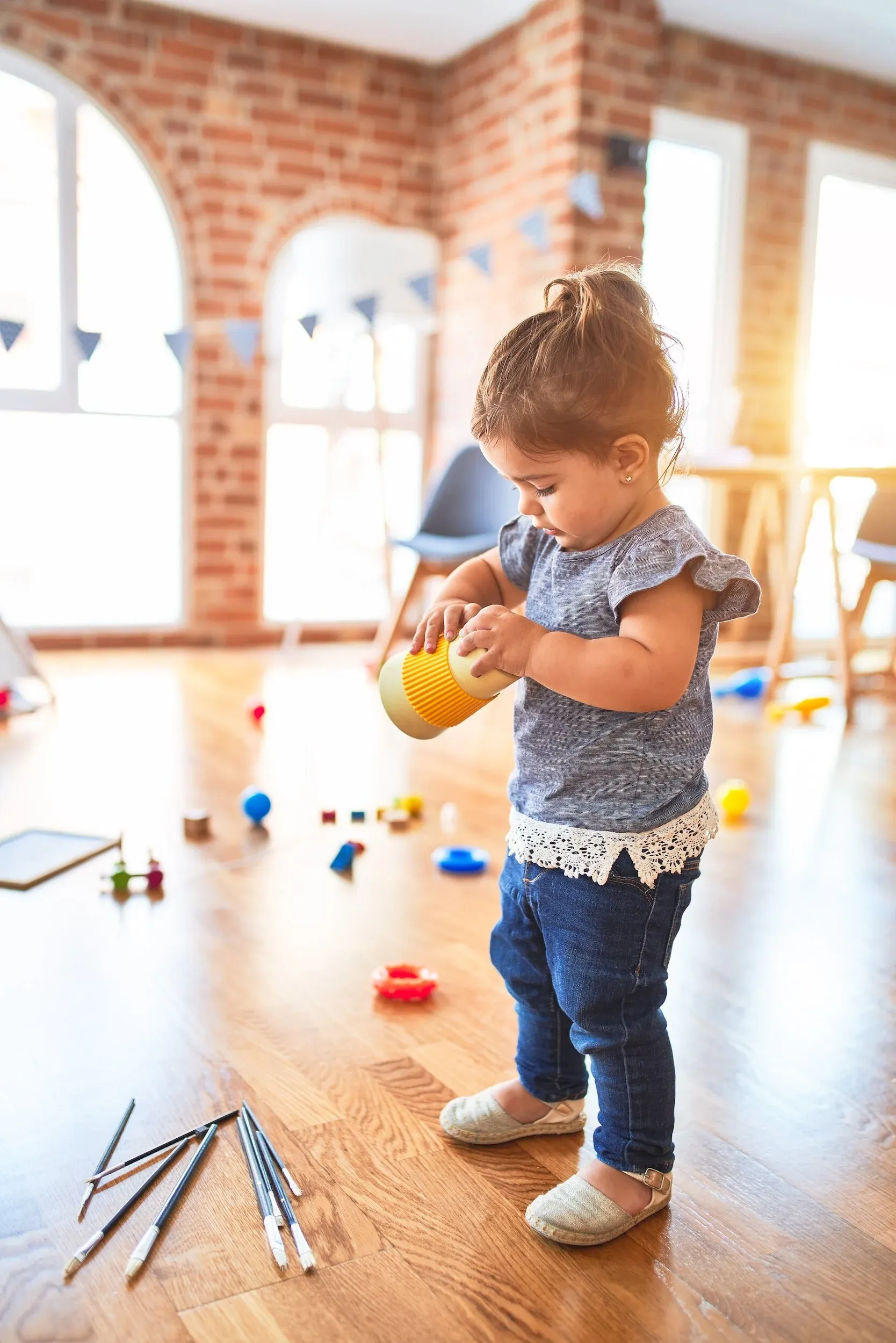 A toddler girl in a bright playroom holding a yellow toy, with blocks scattered on the wooden floor